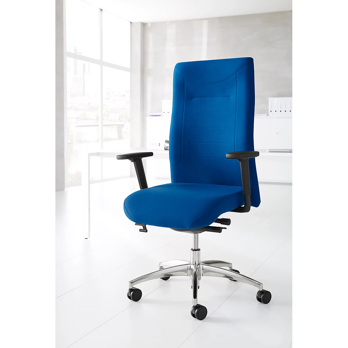 Blue office chair with high back. Black armrests, silver frame with wheels. Background: white office with window.