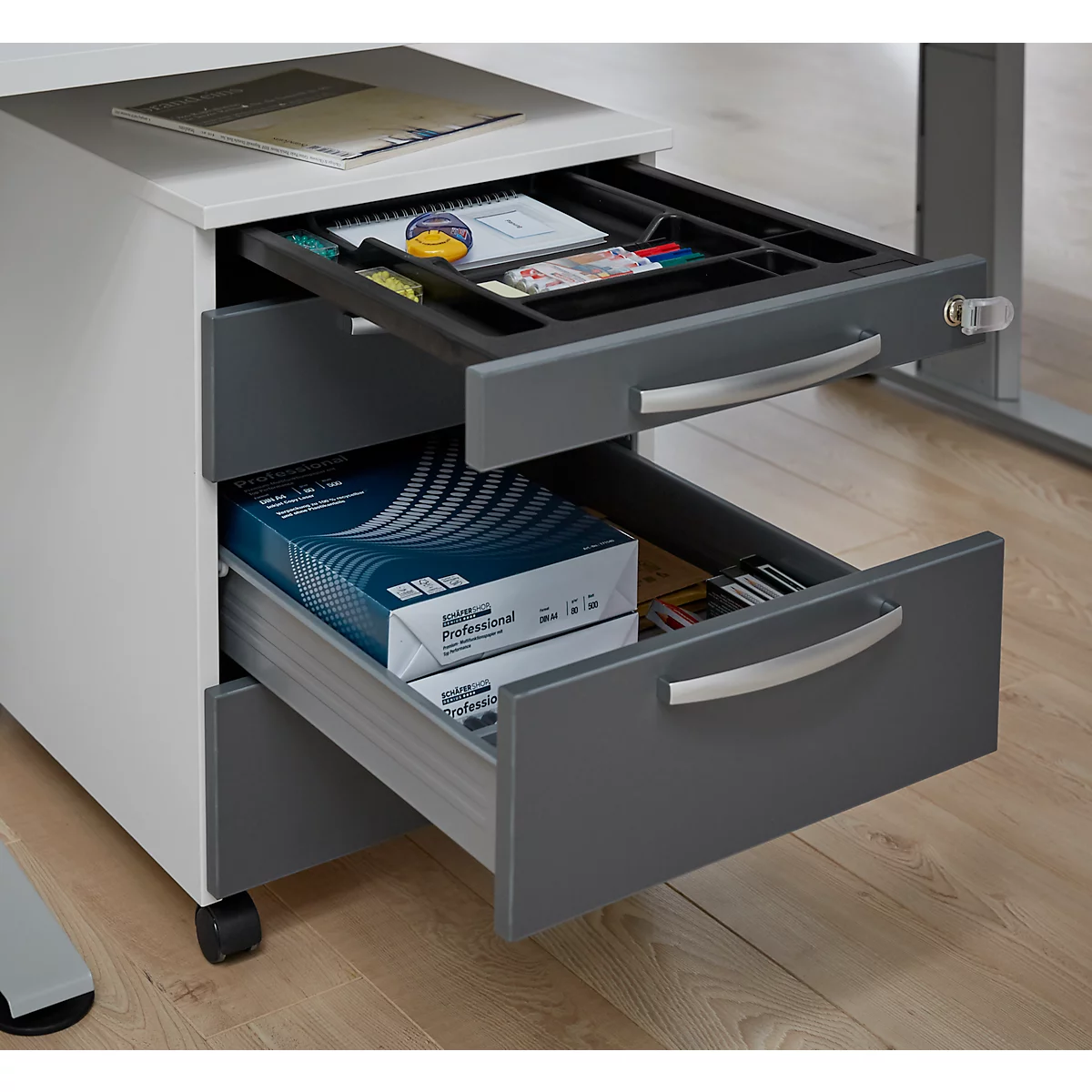 Three drawers of a mobile pedestal, partially opened. The top drawer contains writing utensils, the middle drawer a pad of paper. The bottom drawer is filled with paper.