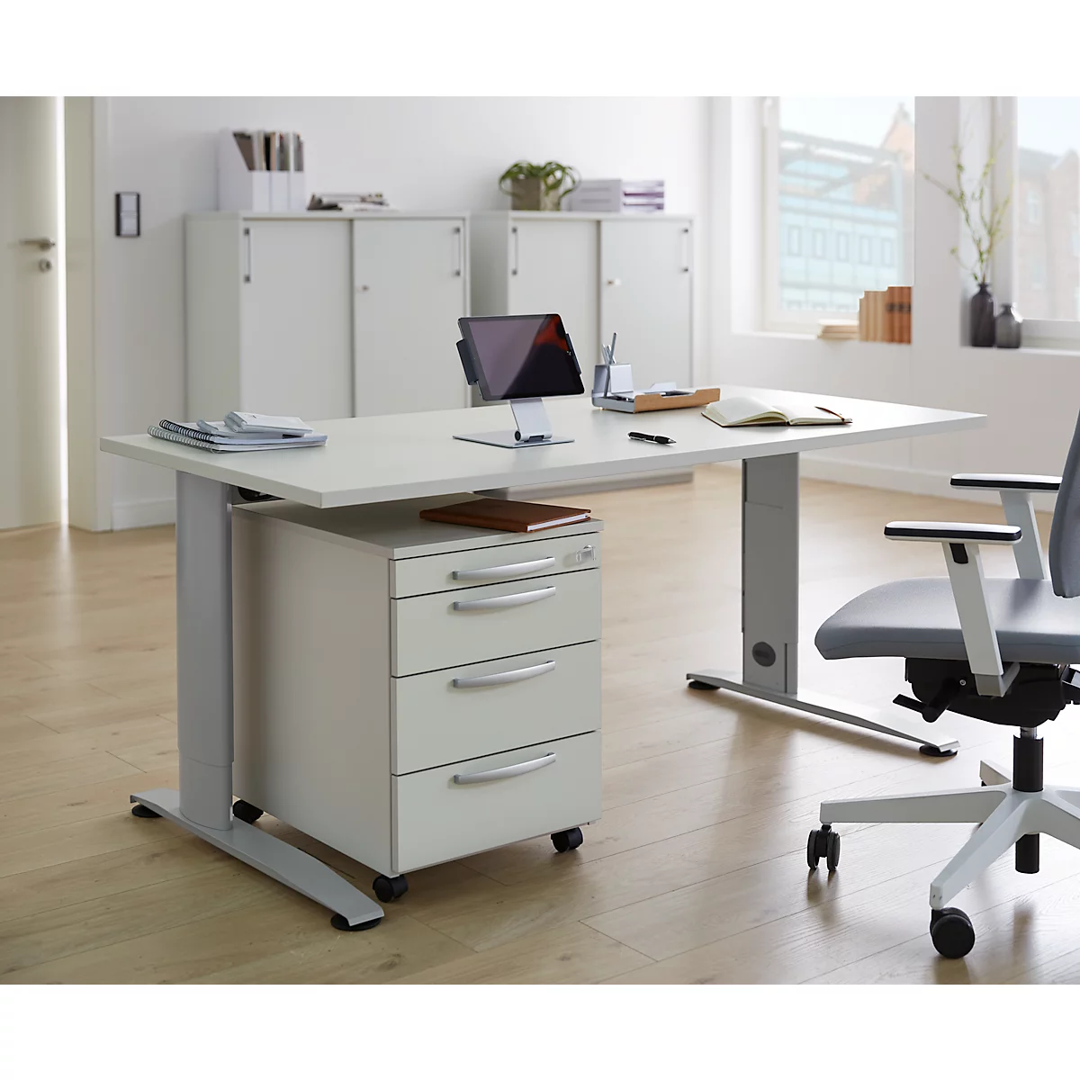 White desk with mobile pedestal, tablet, chair, and window in the background.