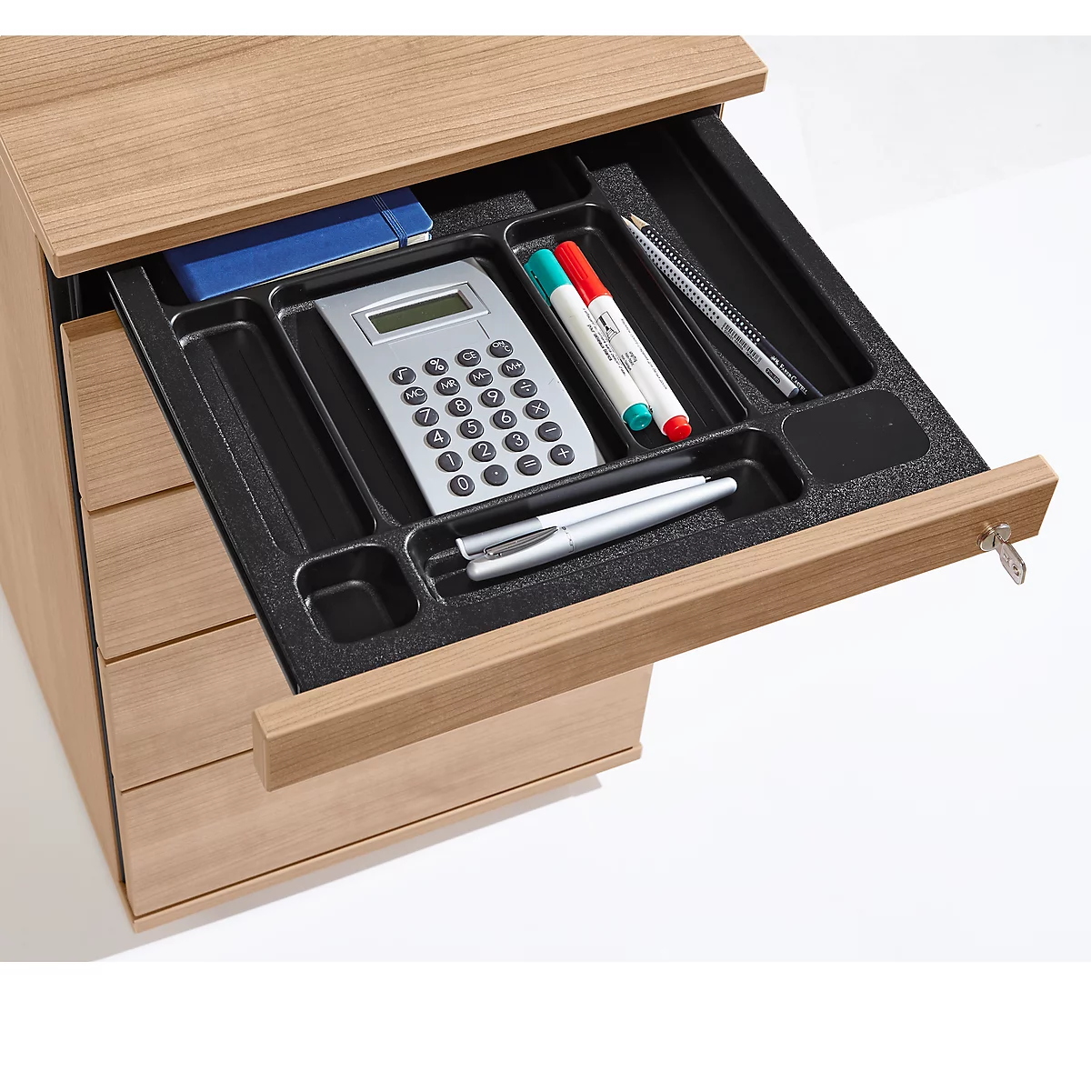An opened wooden desk drawer with black interior. Inside: calculator, pens, notebook and key.