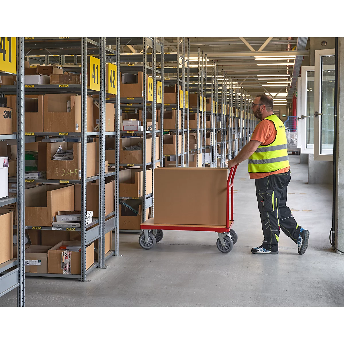 Een man in een veiligheidsvest duwt een kar met een kartonnen doos door een magazijn. Schappen met dozen, gele borden met nummers.