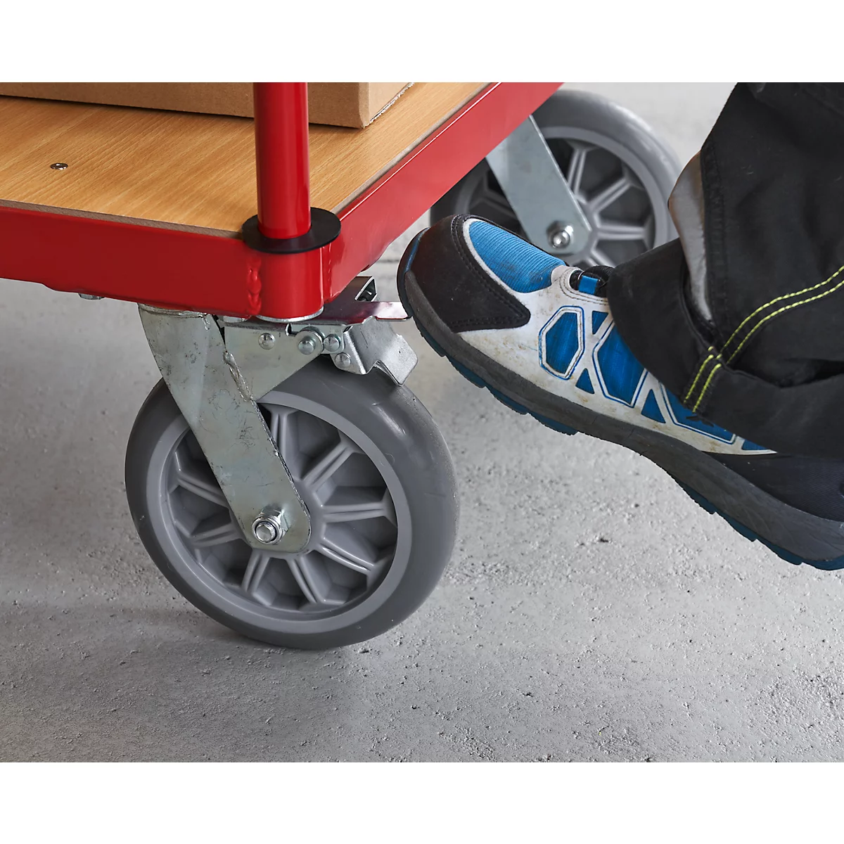 De voet van een man activeert de rem van een rode steekwagen. Het wiel is grijs, het oppervlak is beton.