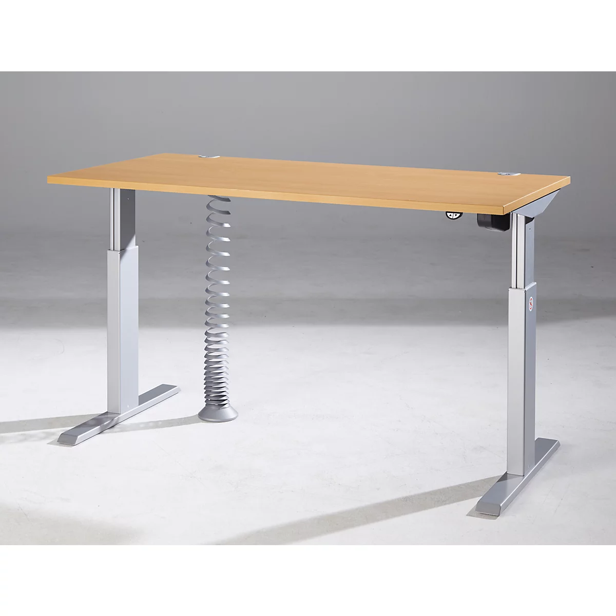 Ein höhenverstellbarer Schreibtisch mit hellbrauner Holzplatte und silbernen Beinen. Eine besondere Federstütze ist sichtbar.