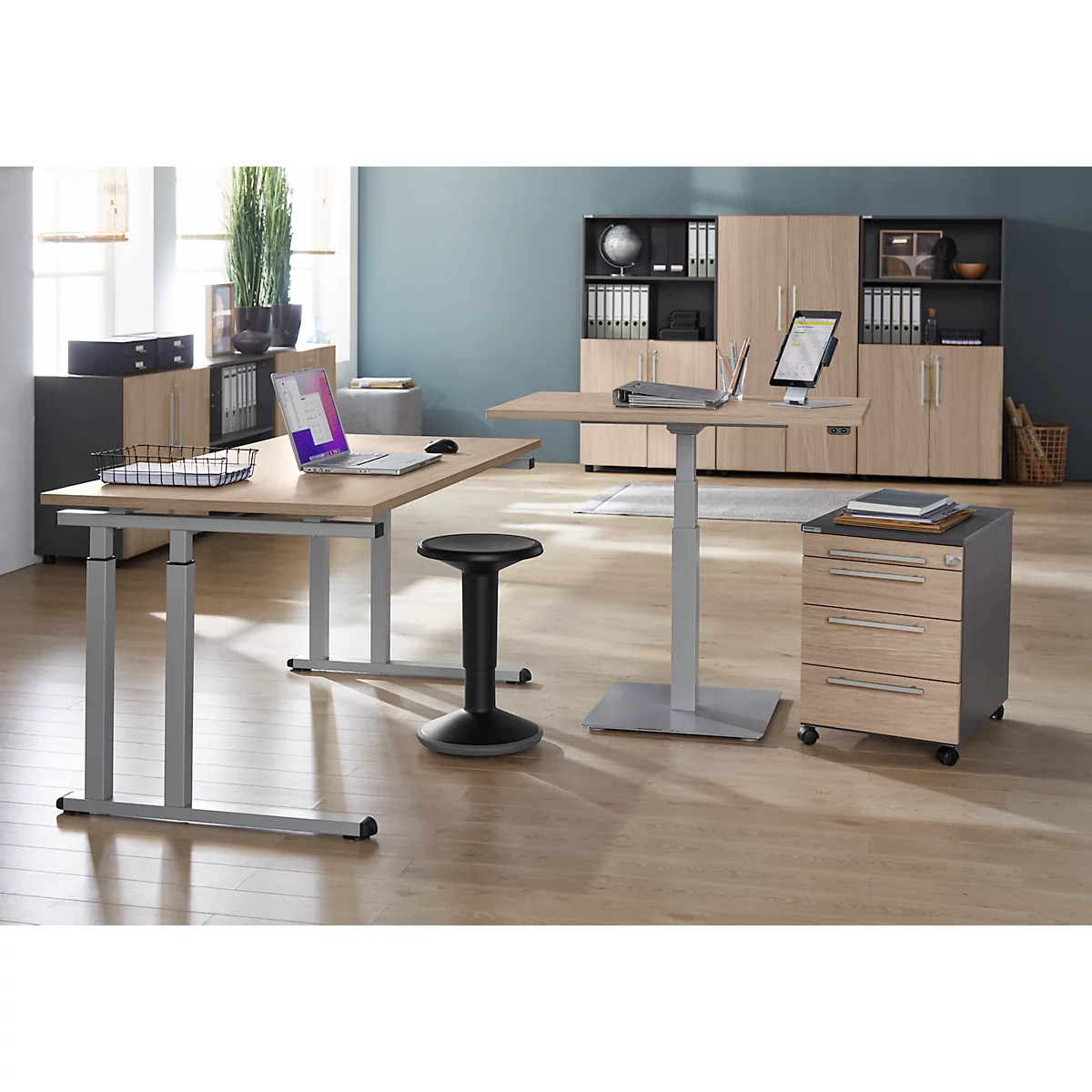 Büro mit zwei höhenverstellbaren Tischen, einem Stehhocker und Aktenschränken. Auf einem Tisch steht ein Laptop und auf dem anderen ein Tablet.