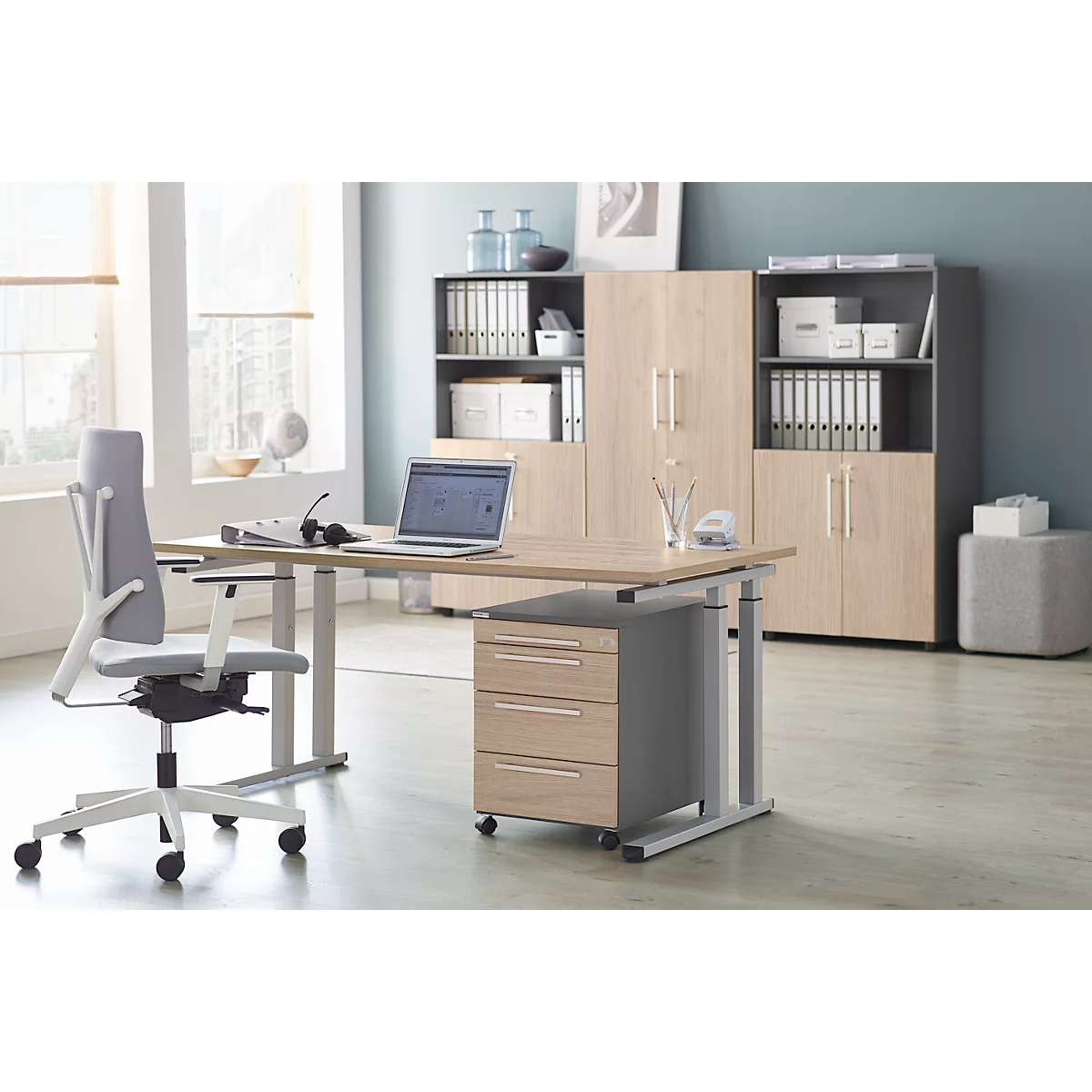 Ein Büro mit höhenverstellbarem Schreibtisch, Drehstuhl und Schrankwand. Auf dem Tisch stehen Laptop und Headset.