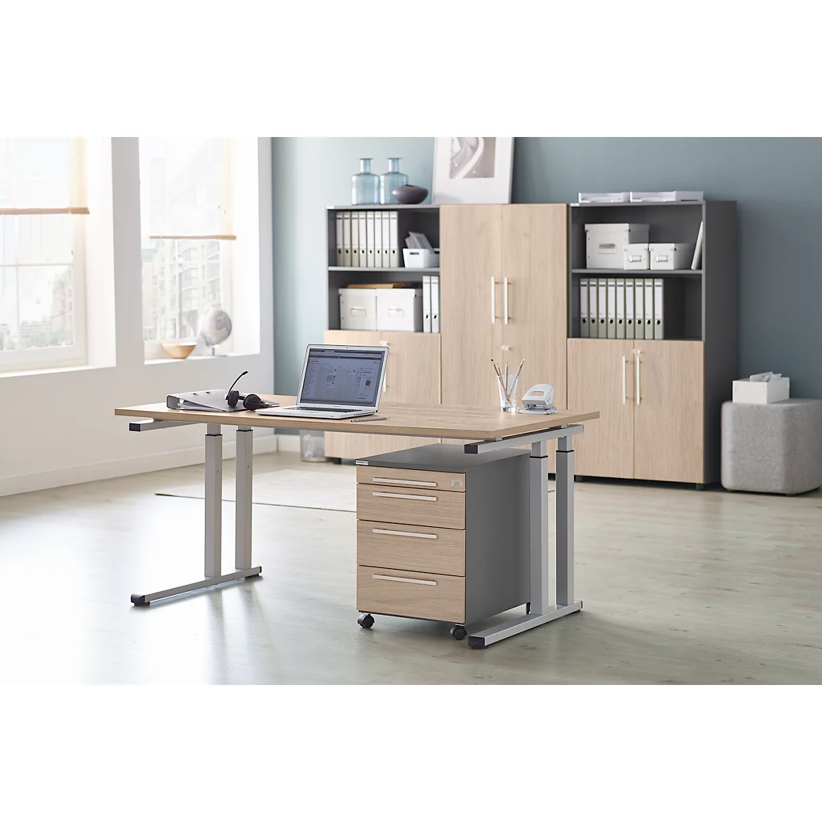 Ein heller Schreibtisch mit Laptop und Rollcontainer. Im Hintergrund Aktenschränke.