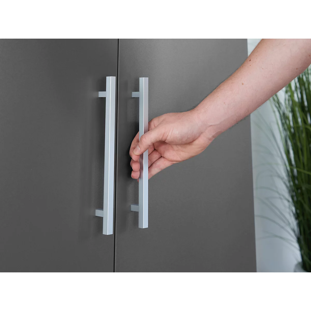 A hand opening a grey cabinet door. The handle is light and elongated. Plants in the background.