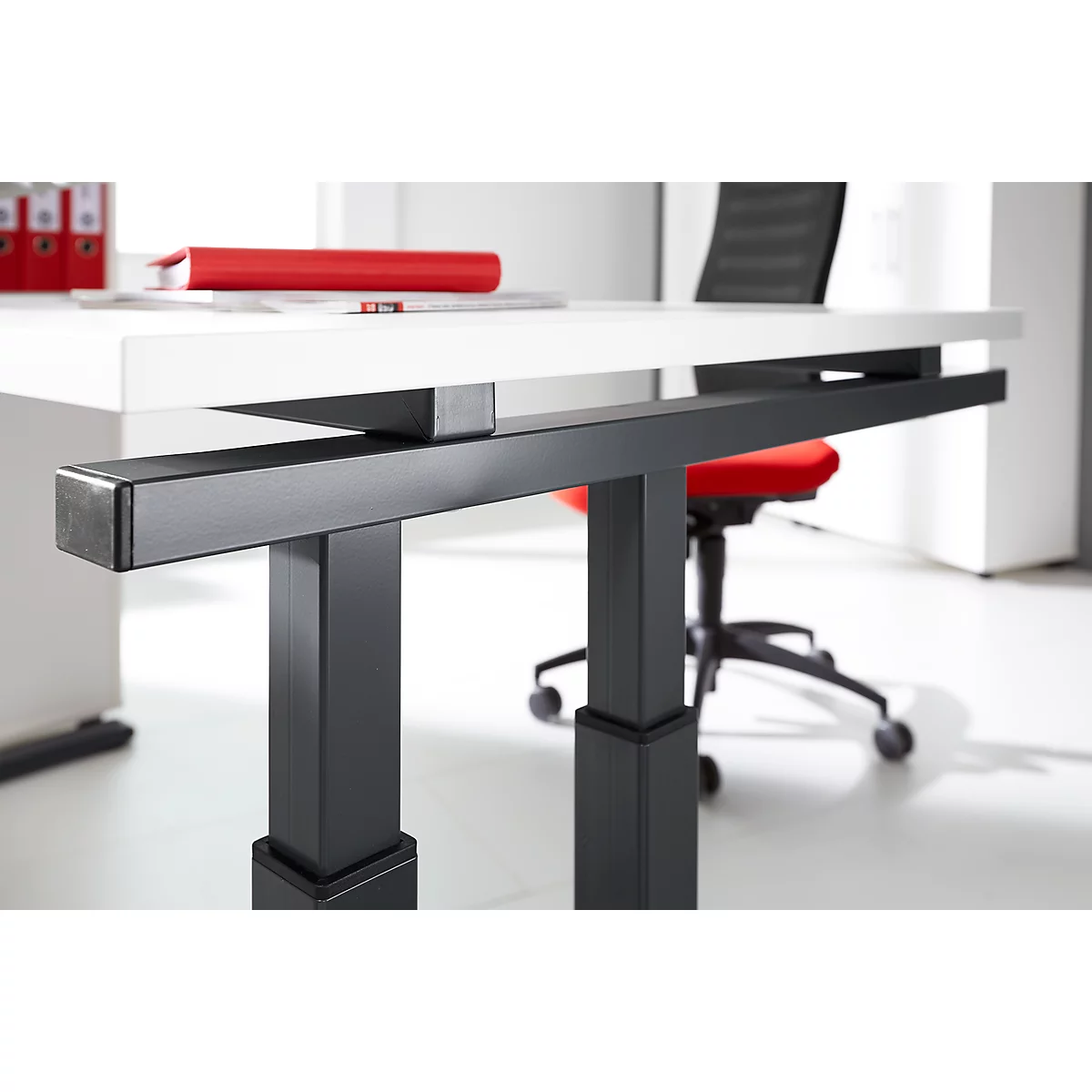 Close-up of the black metal frame of a height-adjustable desk. Desk top white, office chair red.