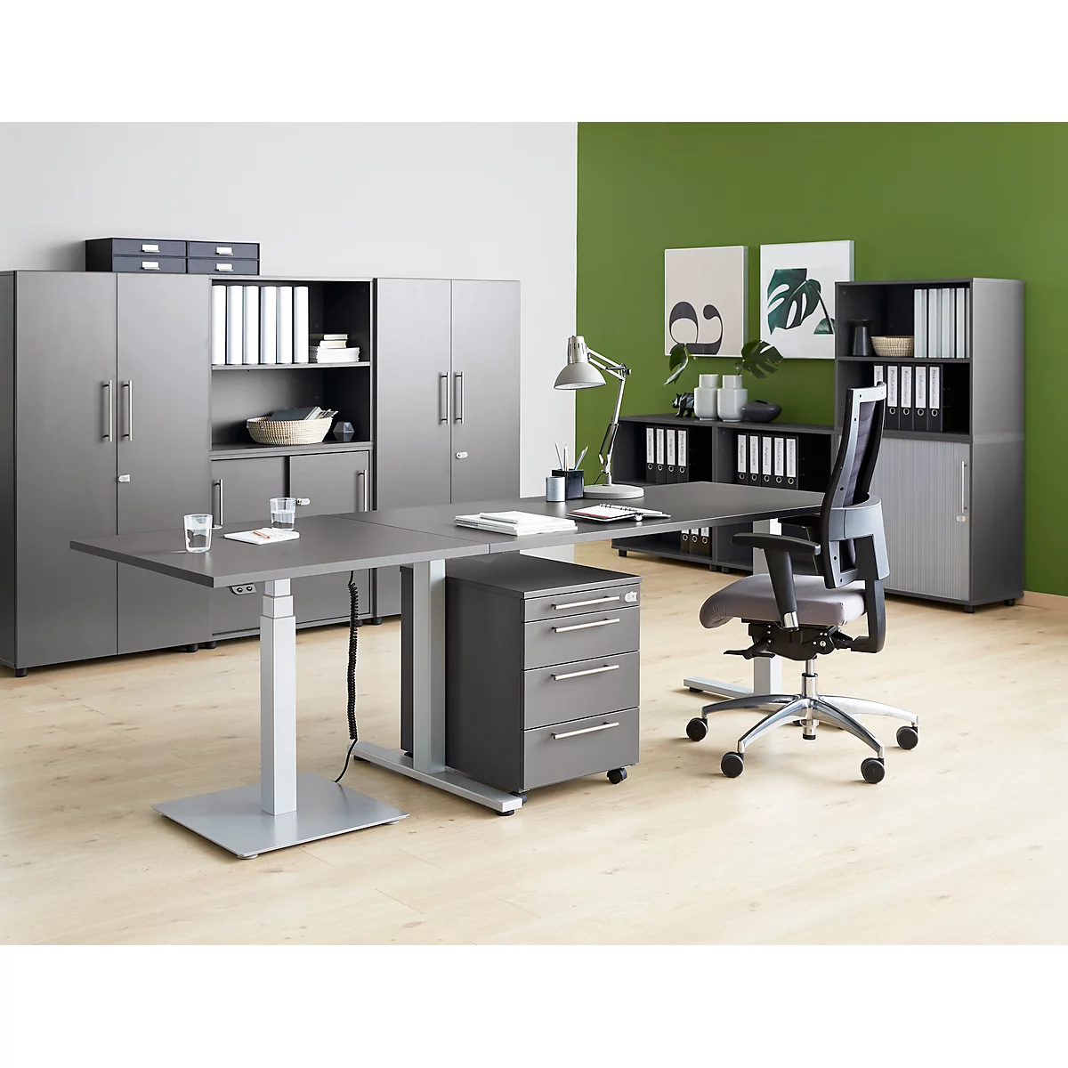 Office with grey furniture and a height-adjustable desk. A laptop, a lamp, office supplies and folders are on the table.