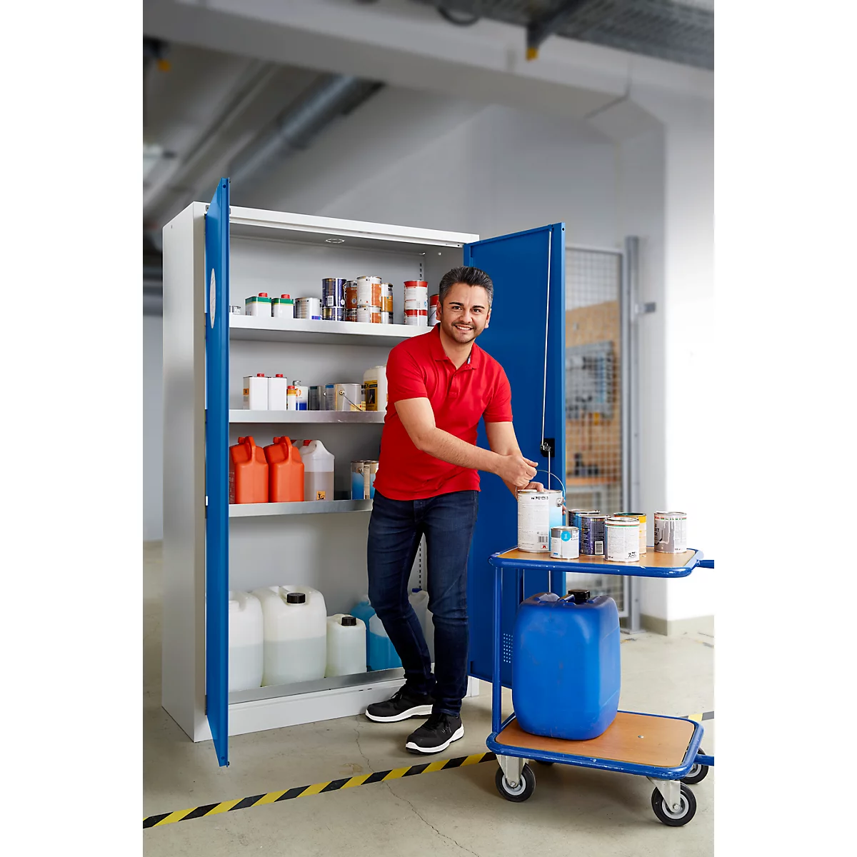 Ein Mann steht vor einem geöffneten Stahlschrank mit Farbeimern und Kanistern, lächelnd in rotem Shirt. Daneben ein Wagen.