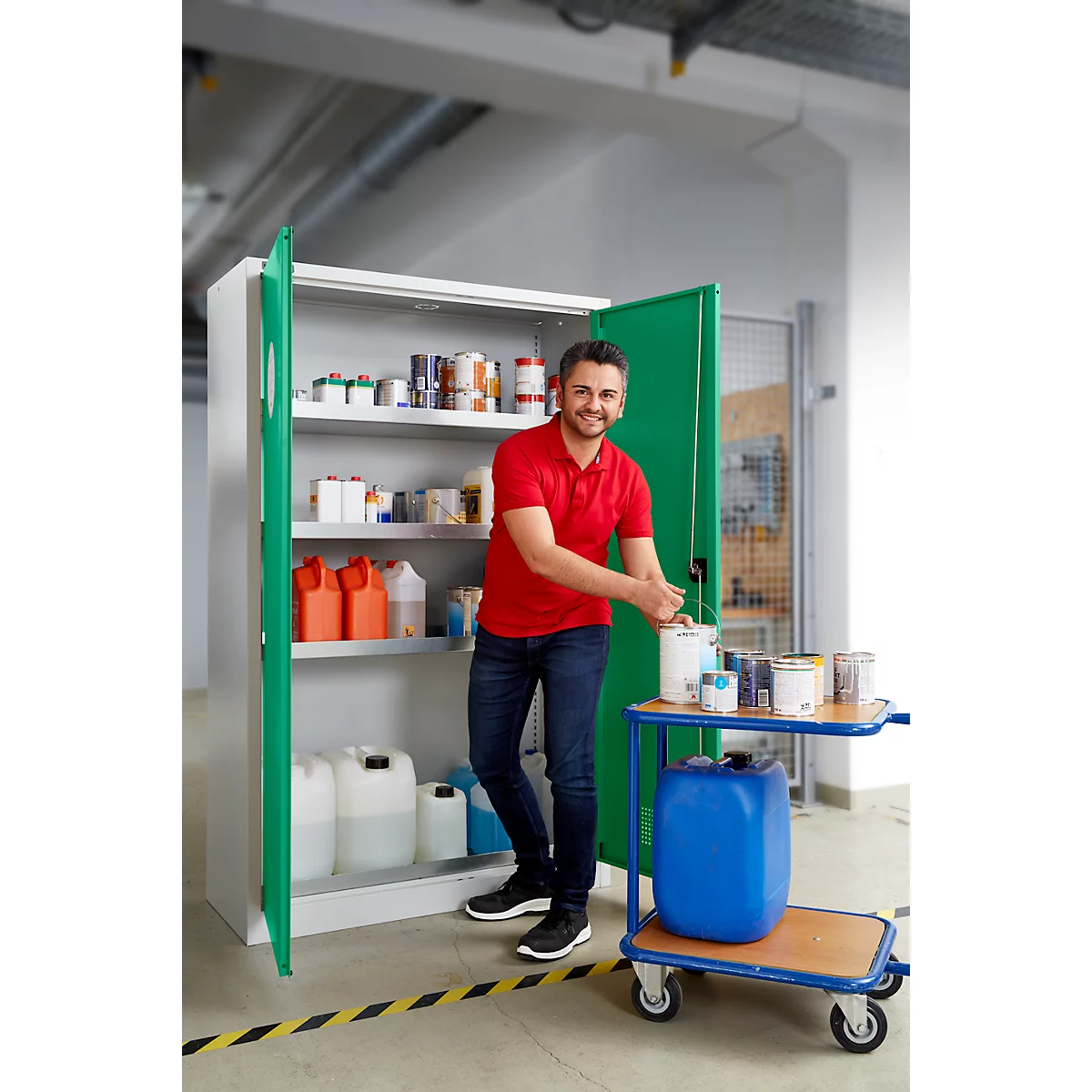 Ein Mann in rotem Shirt steht vor einem Schrank und hält eine Farbe. Der Schrank und ein Wagen sind mit Farben und Behältern gefüllt.