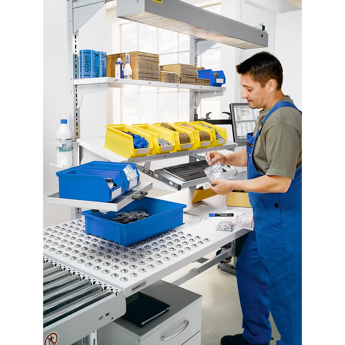 A man in blue workwear sorts parts at an industrial workstation. Shelves with containers and lighting.