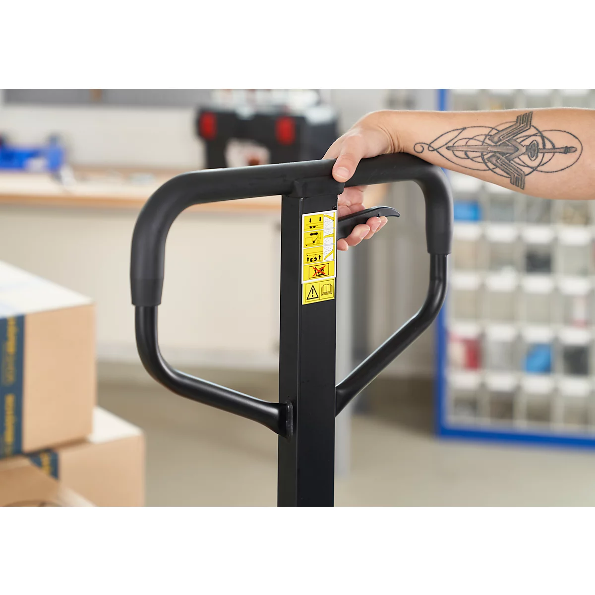 Gros plan d'une main tenant la poignée d'un transpalette noir. Le bras porte un tatouage. Des panneaux d'avertissement jaunes sont fixés au transpalette.