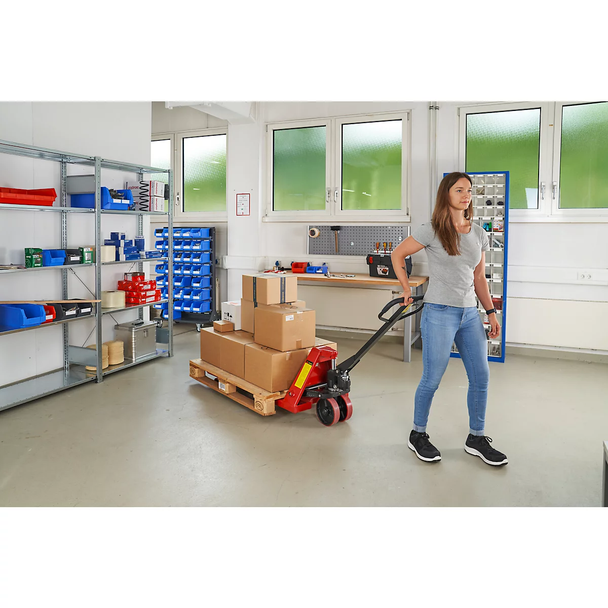 Une femme tire un transpalette avec des cartons dans un atelier. Étagères et fenêtres en arrière-plan.