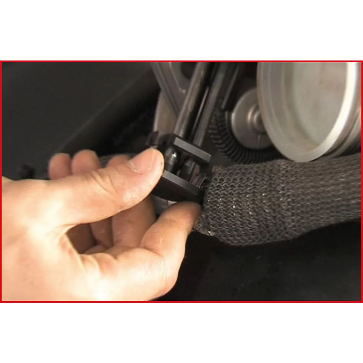 Nahaufnahme einer Hand, die ein schwarzes Kunststoffteil an einem Metallteil befestigt. Im Hintergrund weitere Fahrzeugkomponenten.