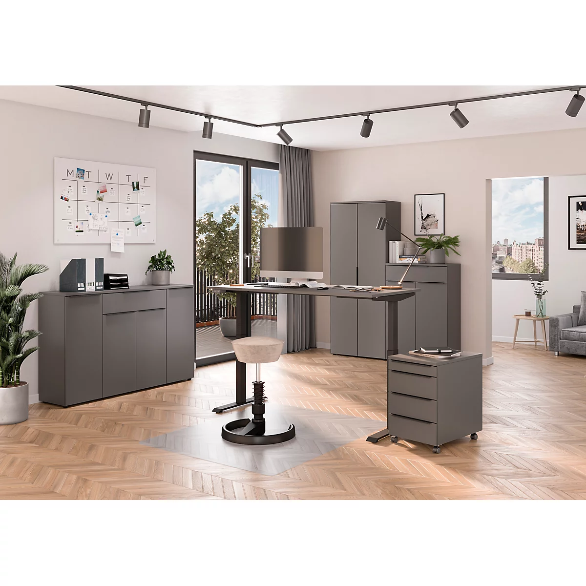 Modernes Heimbüro mit grauen Möbeln, höhenverstellbarem Schreibtisch, Rollcontainer und hellem Stehhocker auf Parkettboden.