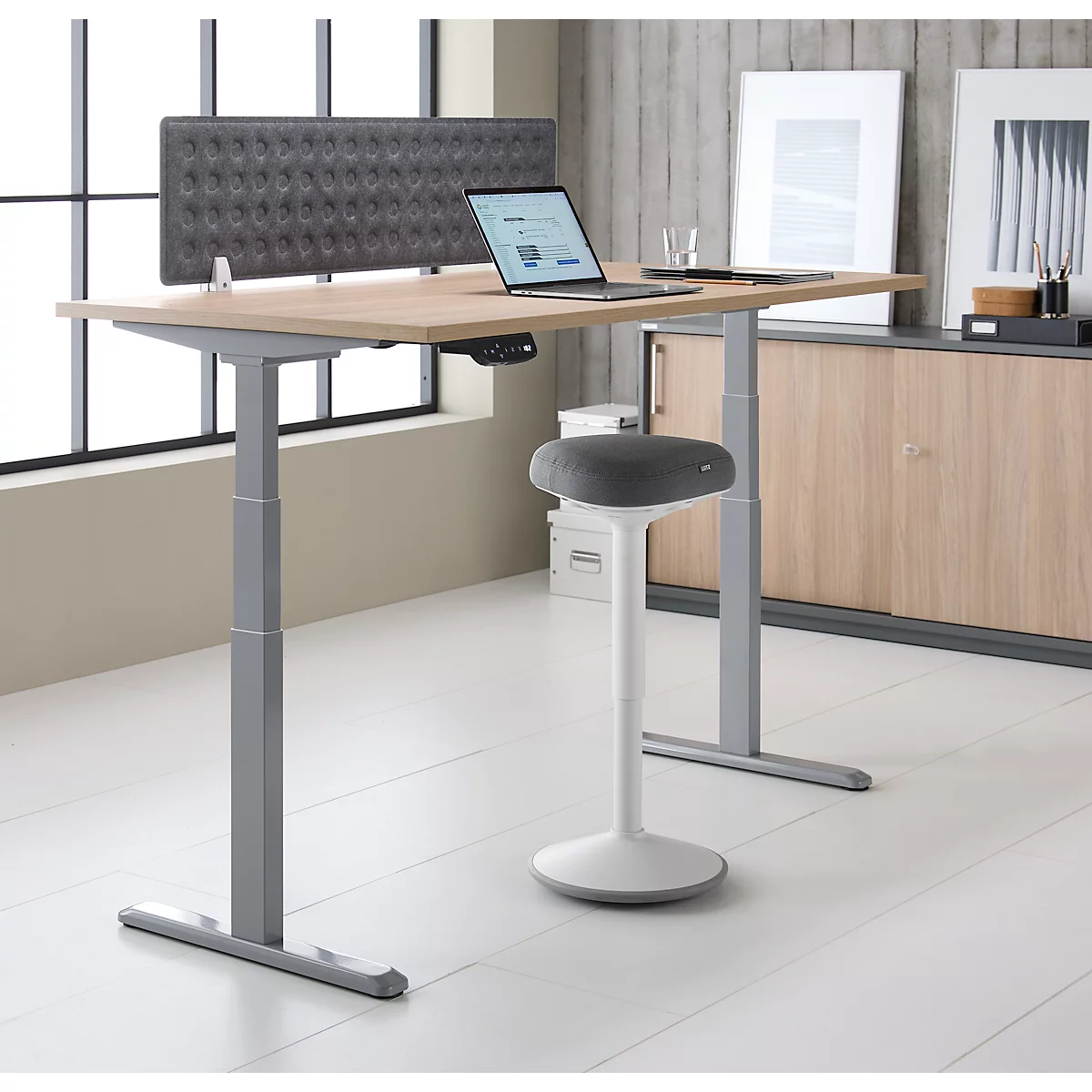 Heller Schreibtisch mit höhenverstellbarem Gestell, Laptop und Hocker; im Hintergrund graue Wand.