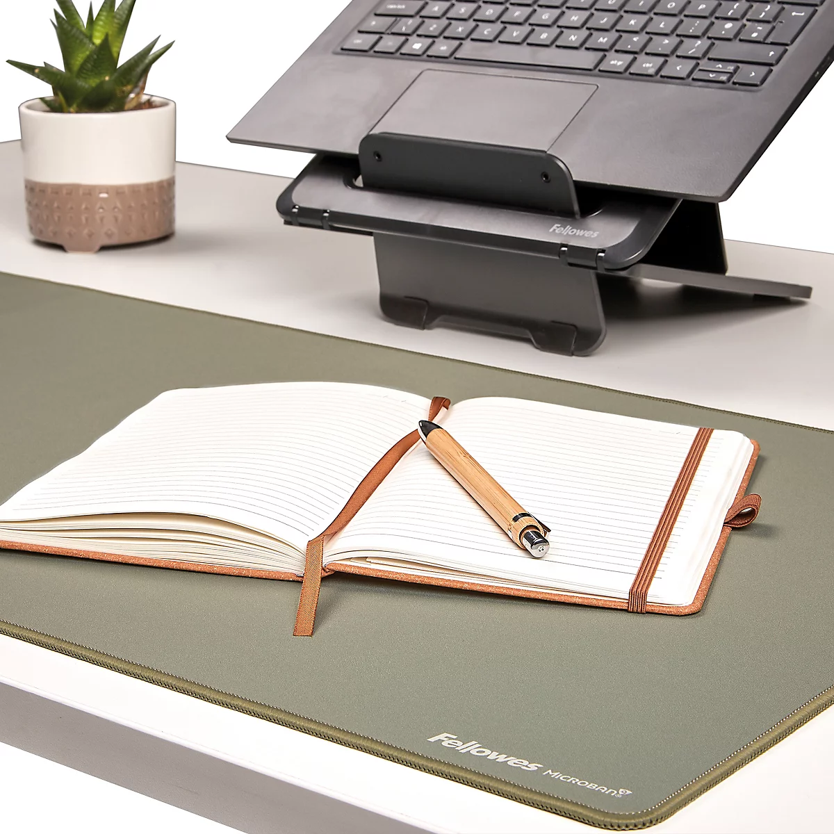 Schwarzer Laptop auf Ständer, darunter Tastatur, Maus, Notizbuch mit Stift und Pflanze. Vor weißem Hintergrund.
