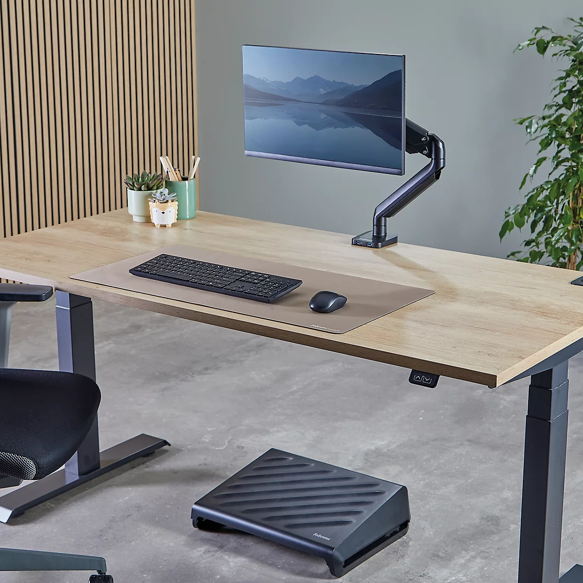 Schreibtisch mit Monitor, Keyboard, Maus, Notizbuch und Topfpflanze. Monitor zeigt Landschaft. Graue Unterlage für das Zubehör.