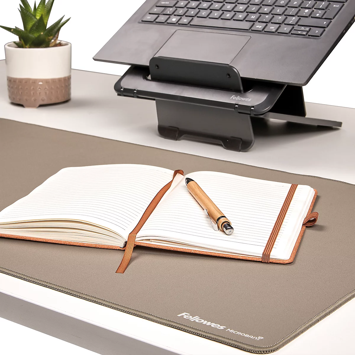 Schwarzer Laptop auf Ständer. Darunter Maus, Tastatur und Notizbuch. Kleiner Topf mit Pflanze.
