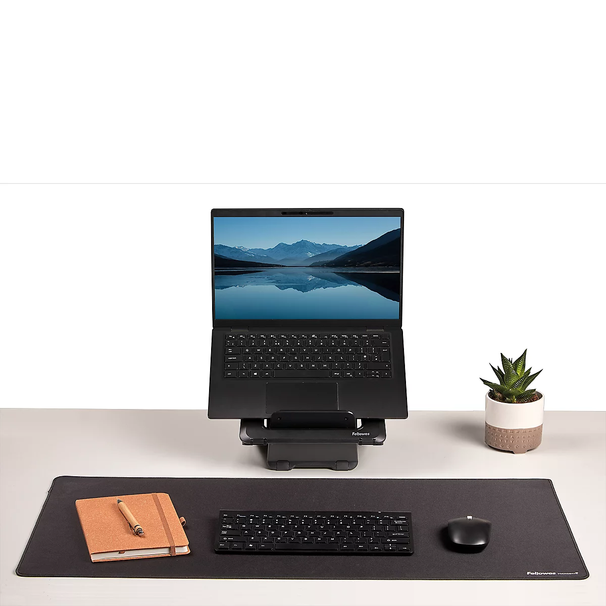 Schwarzer Laptop auf einem Ständer, umgeben von Zubehör: Maus, Tastatur, Notizbuch, Topfpflanze.