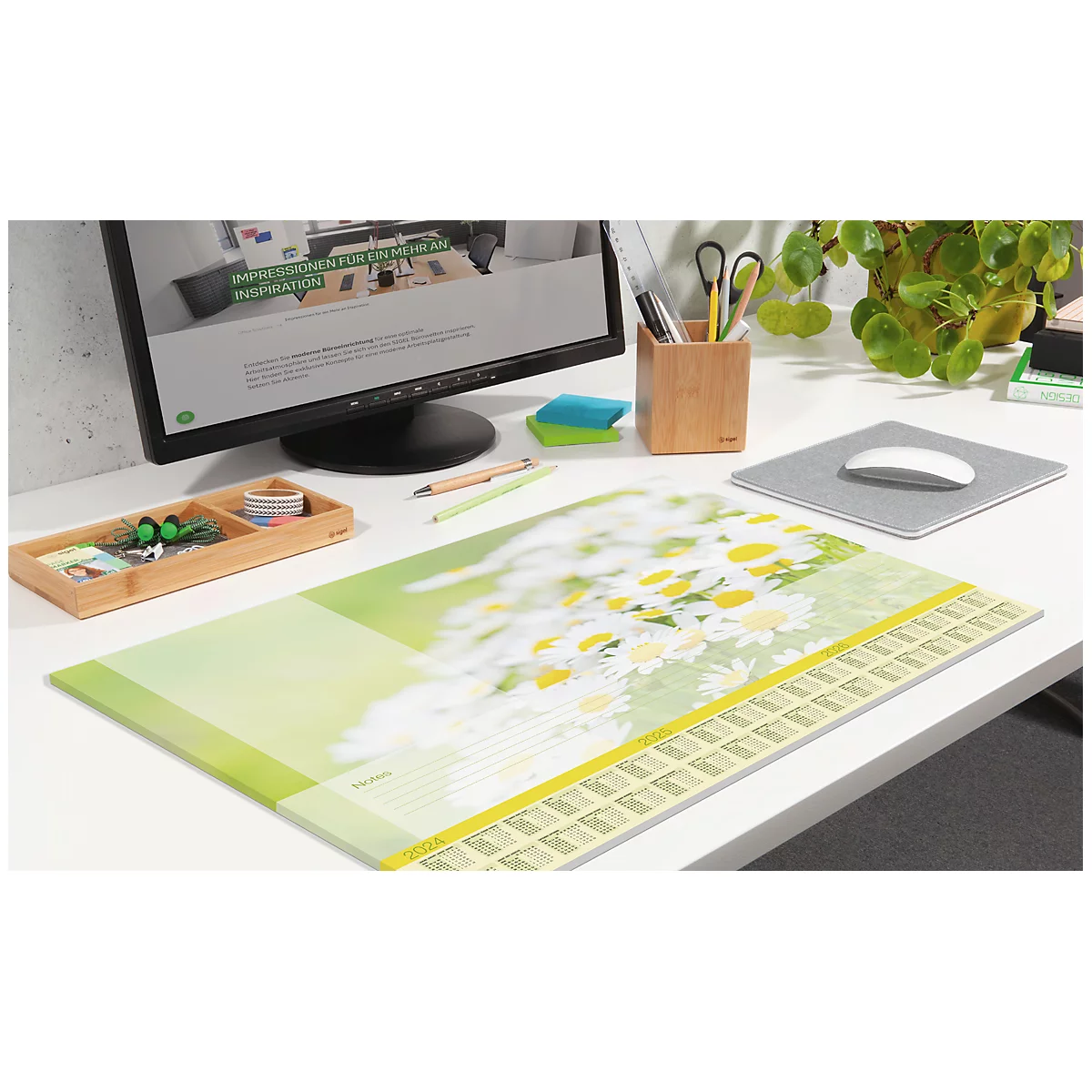 Schreibtisch mit Mauspad, Kalender und Zubehör. Im Hintergrund ein Monitor und eine Pflanze.