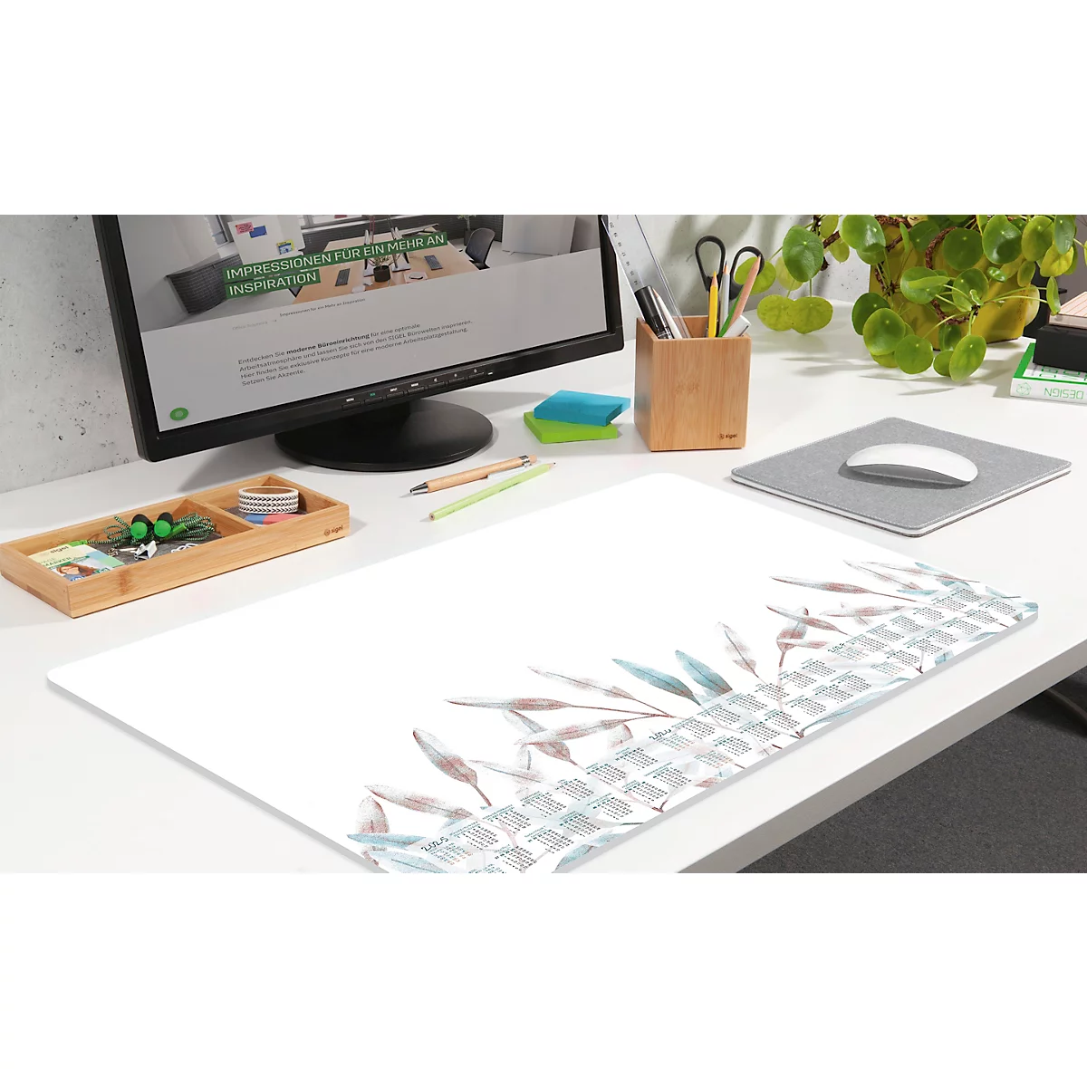 Schreibtisch mit Monitro, Mauspad mit Pflanzen-Design. Im Hintergrund Stiftehalter, Grünpflanze und Büroaccessoires.