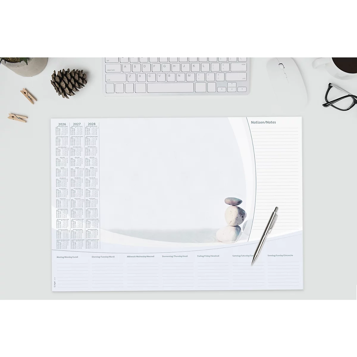 Weisser Schreibtischunterlage mit Steinen, Stift, Kalender und Notizen. Oben links ein Topf, Wäscheklammern, Tannenzapfen und Tastatur.