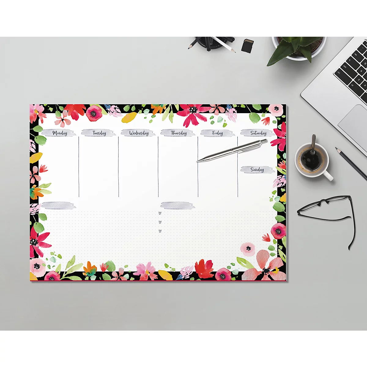 Ein floraler Wochenplaner liegt auf einem Schreibtisch, umgeben von Stiften und Laptop.