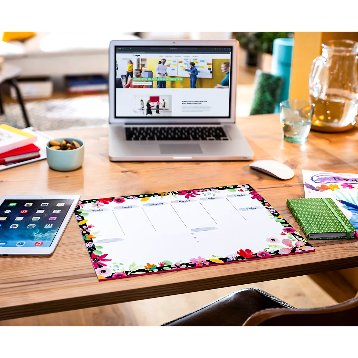 Schreibtisch mit Laptop, Tablet, Notizblock und Wochenplaner mit Blumendekor. Auf dem Laptop ist eine Website zu sehen.