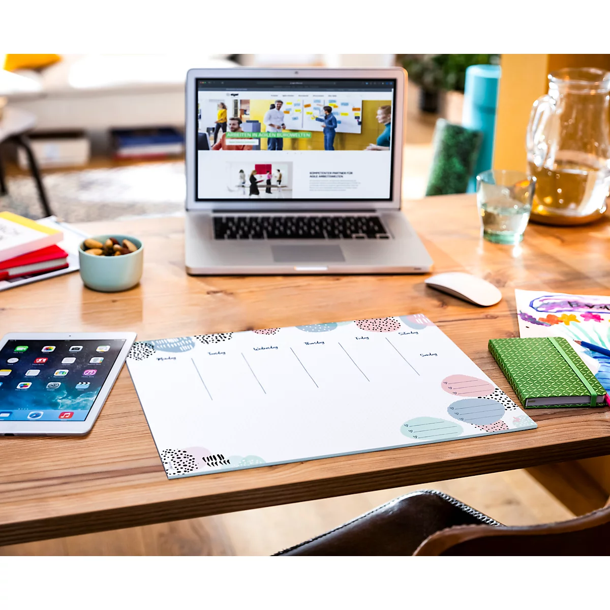 Schreibtisch mit Laptop, Tablet, Notizbuch und Wochenplaner. Laptop zeigt Webseite. Wochenplaner mit bunten Kreisen, Notizbuch grün.
