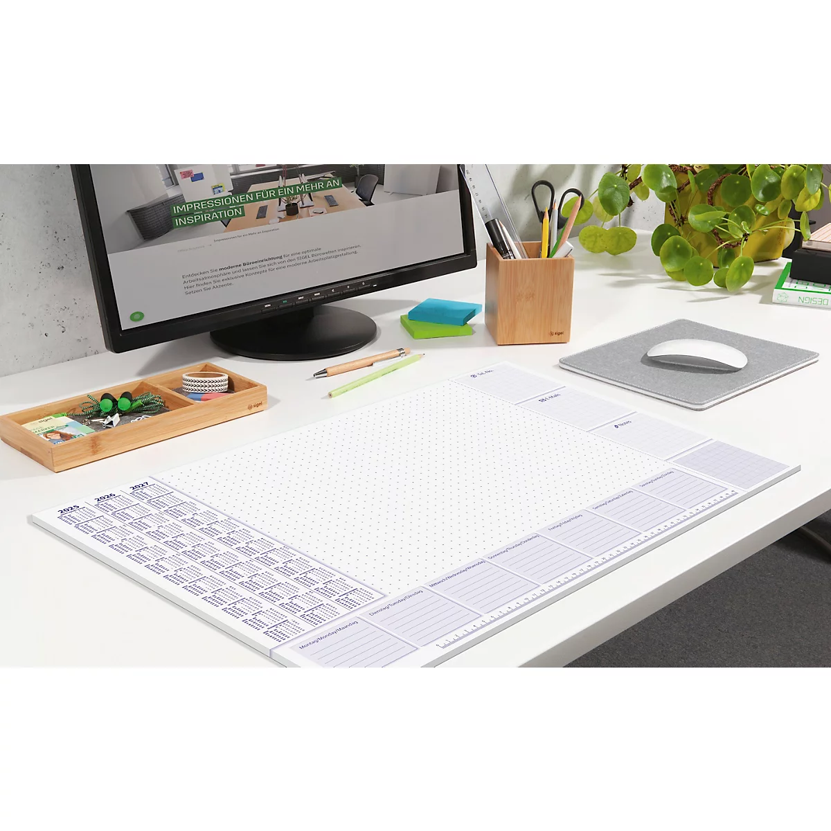 Schreibtisch mit Arbeitsfläche, PC, Stifthalter und Pflanzen. Auf der Arbeitsfläche liegt eine Schreibunterlage mit Kalender.
