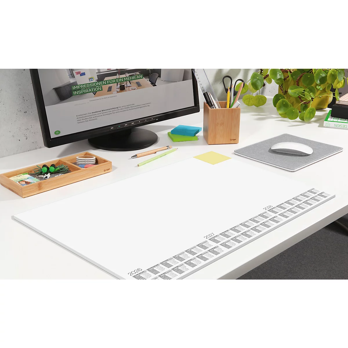 Schreibtisch mit Zubehör: Monitor, Maus, Stifte, Notizzettel und eine weisse, längliche Schreibunterlage mit grauer Markierung.