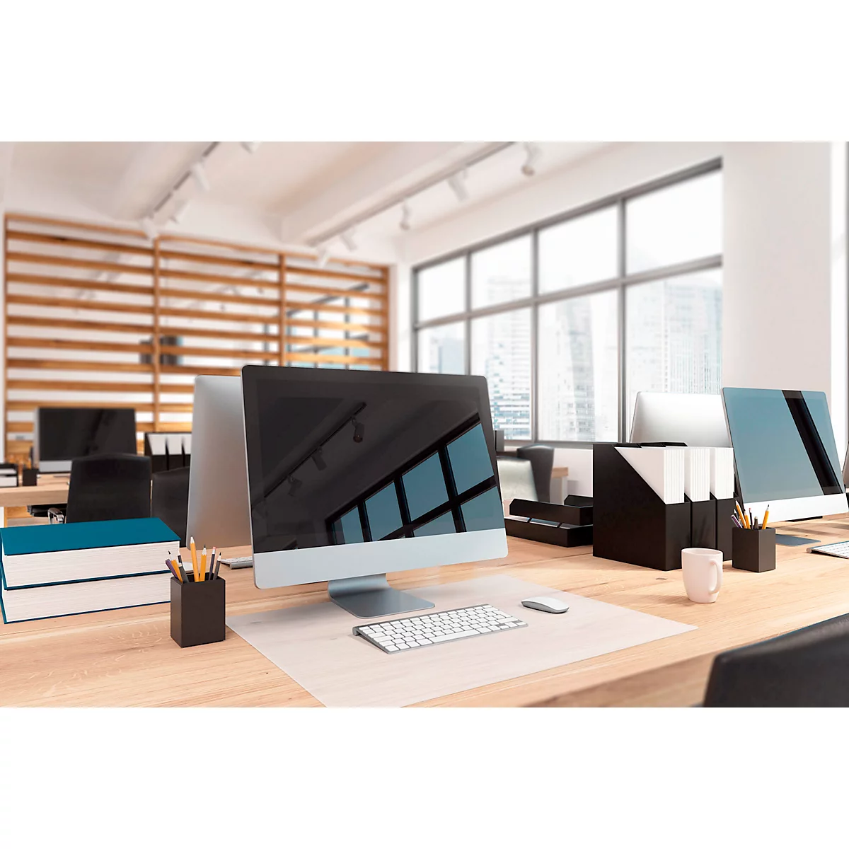 Lichte kantoorruimte met computers op houten tafels. Ramen met uitzicht op gebouwen. Bureau met toetsenbord, muis, pennen, mok.