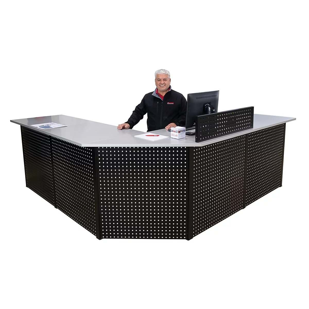 Een man staat achter een zwart receptiebalie met een geperforeerd patroon. Hij glimlacht vriendelijk. Er liggen papier en een computer op de balie.