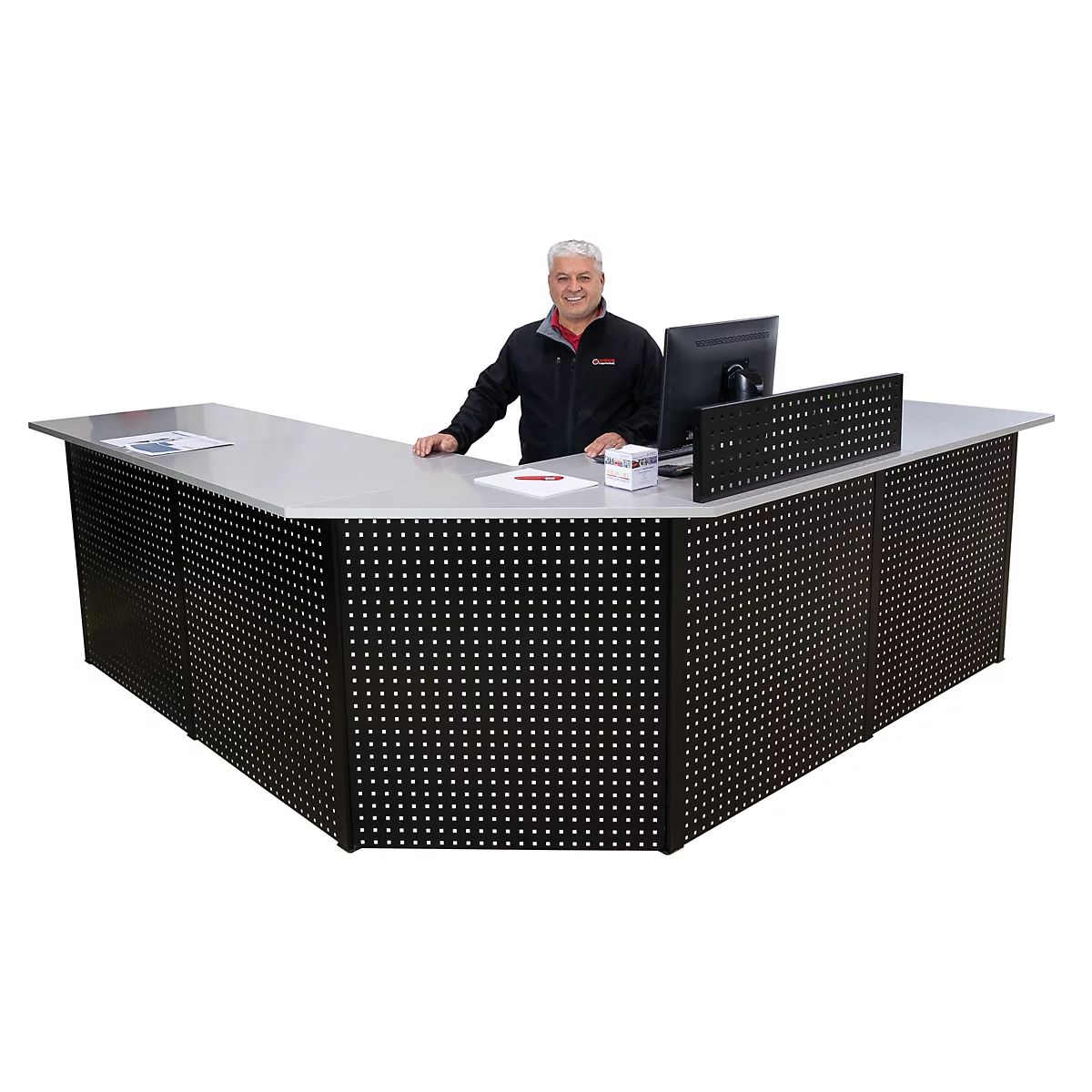 Een man staat achter een zwart receptiebalie met een gaatjespatroon, waarop een computer staat. Hij glimlacht.