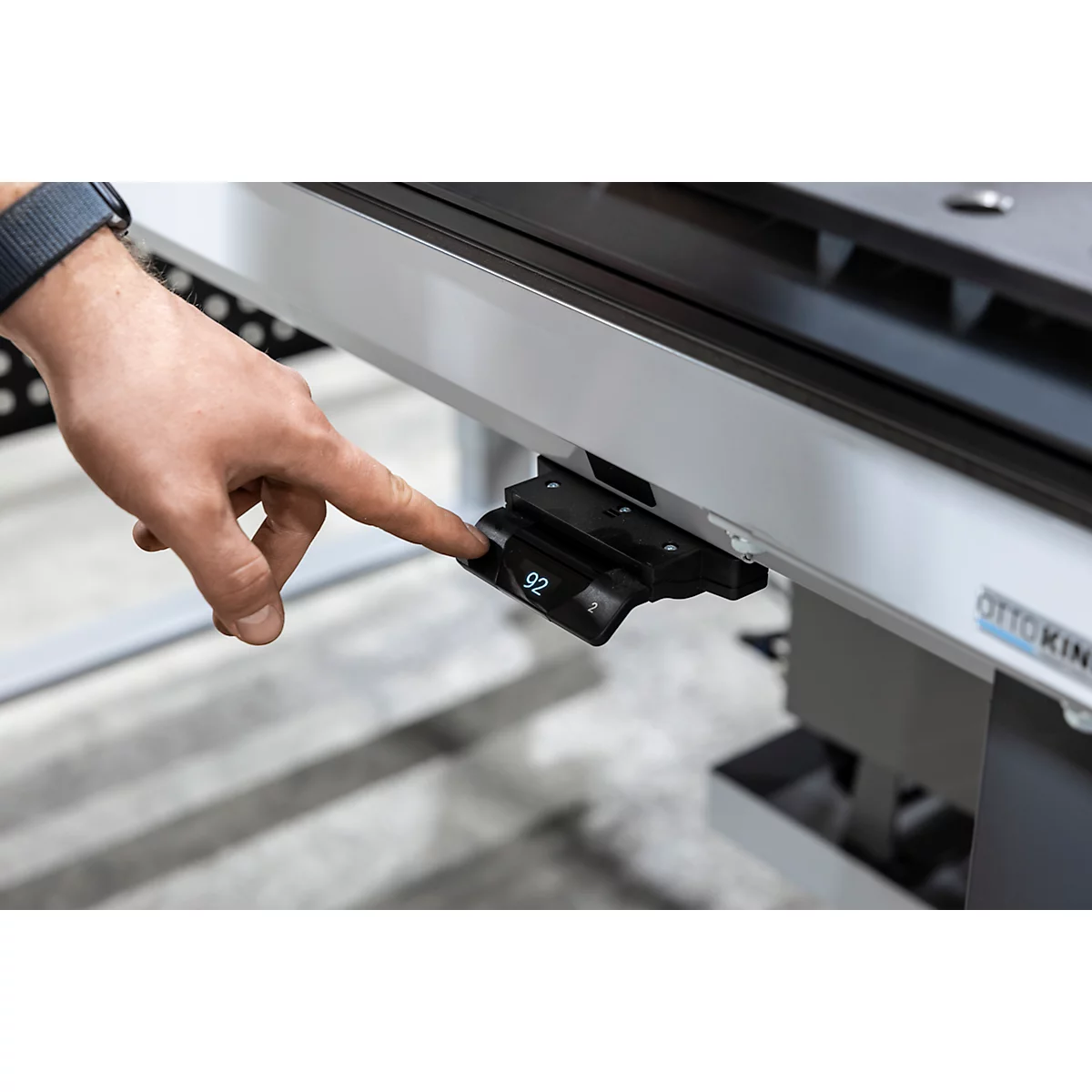 Nahaufnahme einer Hand, die auf ein Bedienfeld drückt, das an einem Schreibtisch befestigt ist. Auf dem Display ist die Zahl 92 zu sehen.