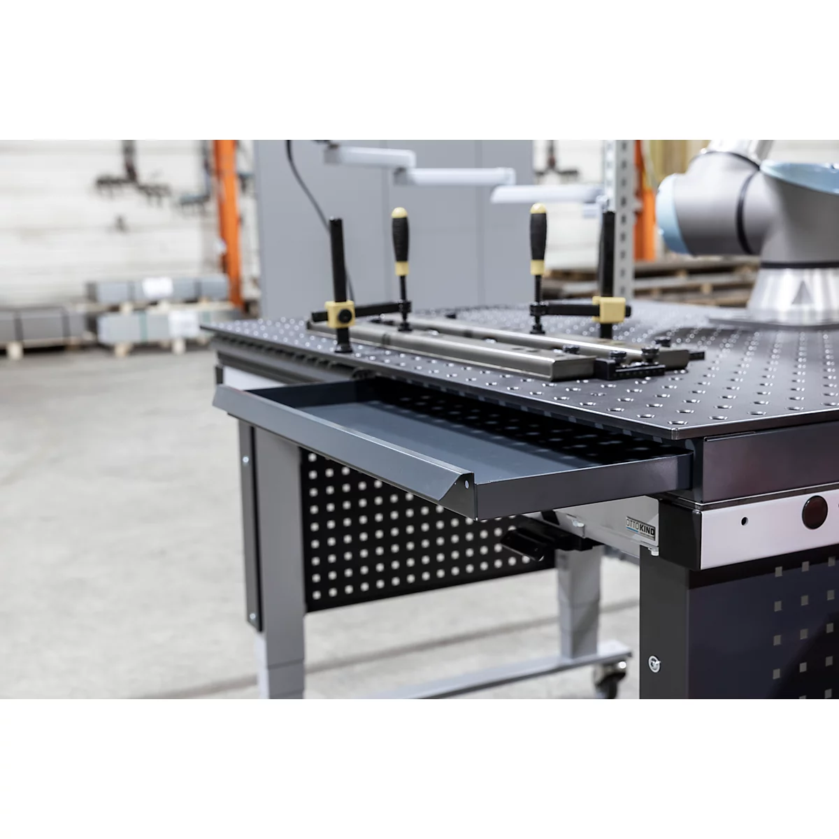 Grauer Werkzeugtisch mit Lochplatte, Schublade, Werkstücken und Schraubzwingen. Im Hintergrund ein Roboterarm.