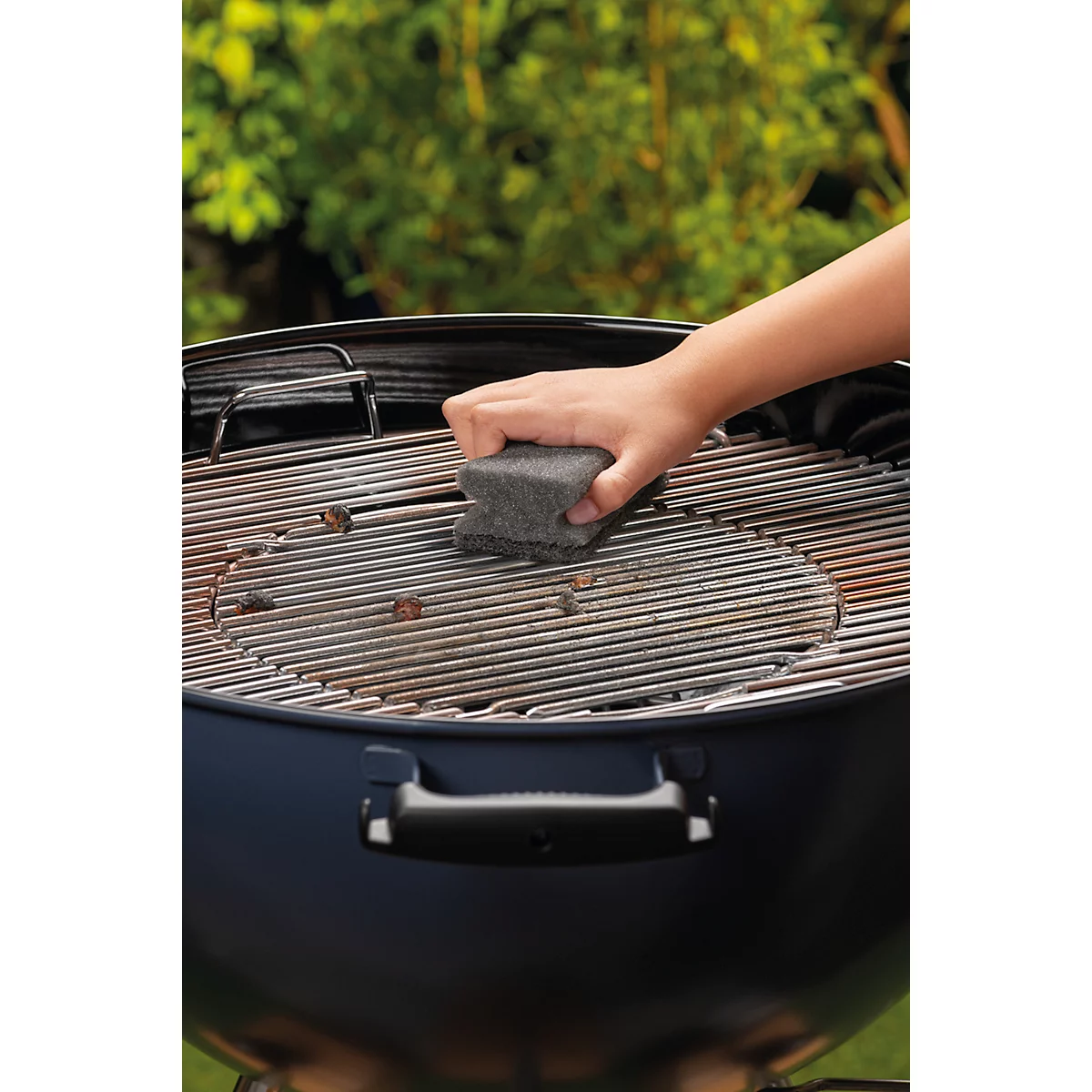 Een hand reinigt de grillrooster van een zwarte grill met een spons.