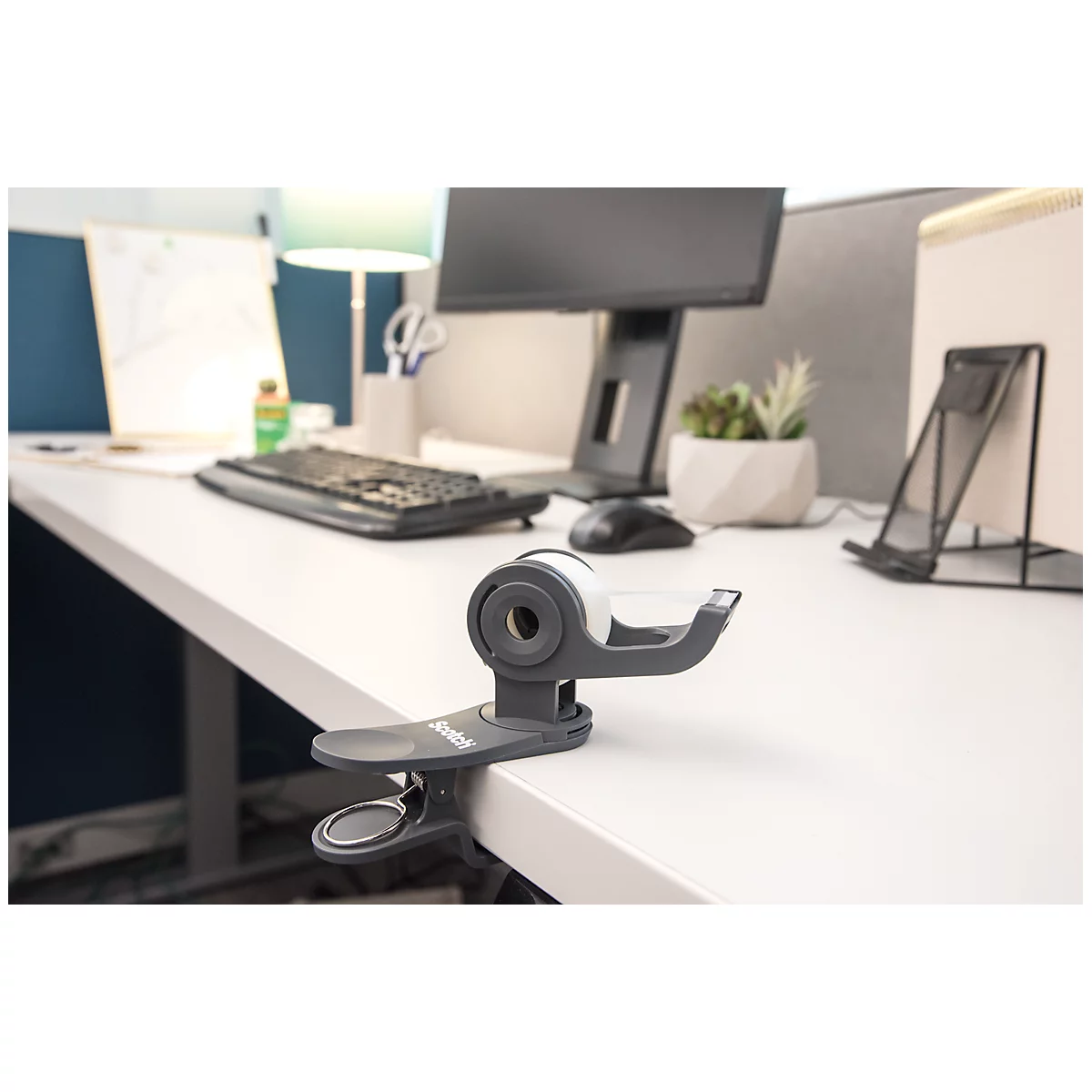 A clip-on tape dispenser on a desk. Background includes a computer, keyboard, desk lamp, and office accessories.