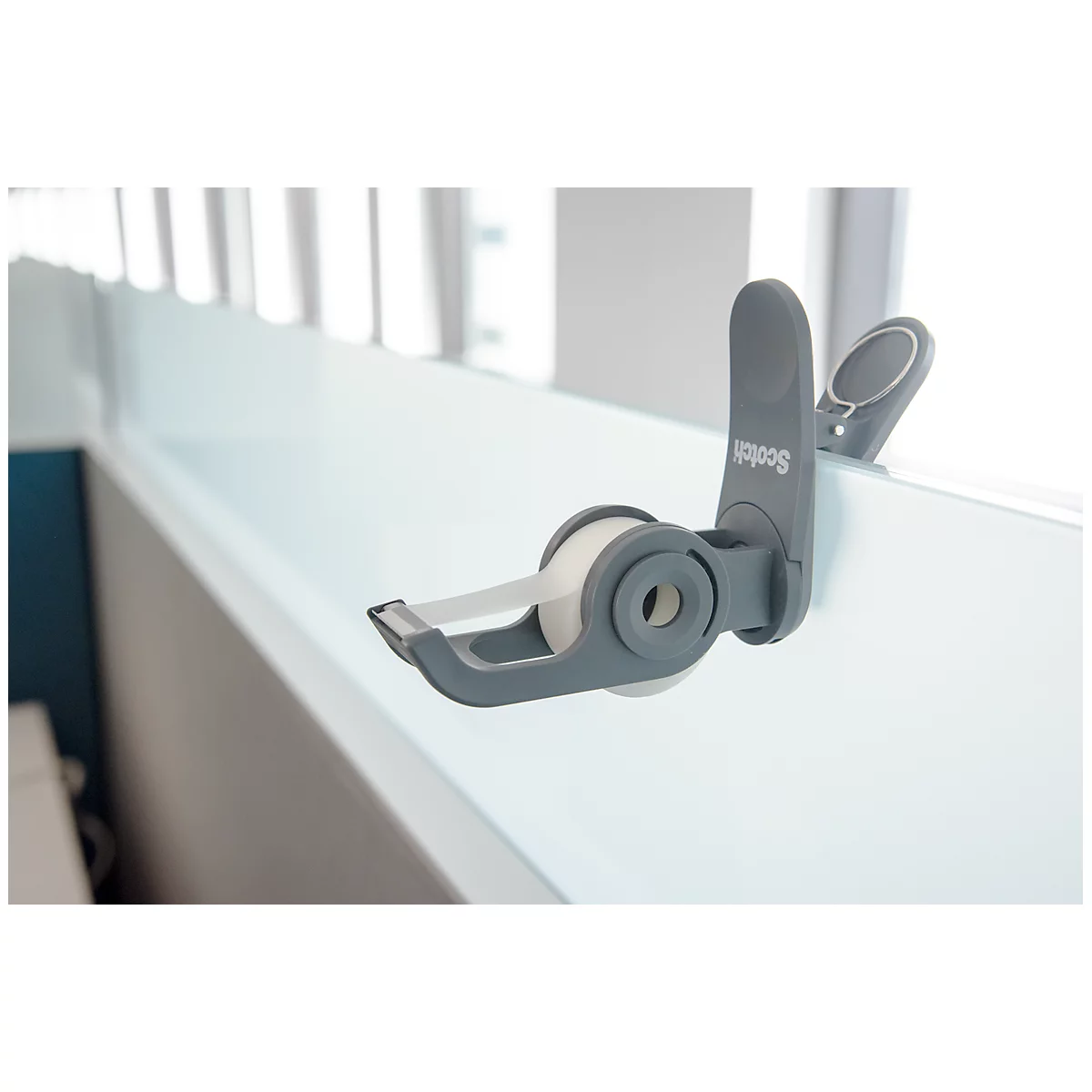 A gray scotch tape dispenser sticks to a white shelf. Transparent tape is being pulled.