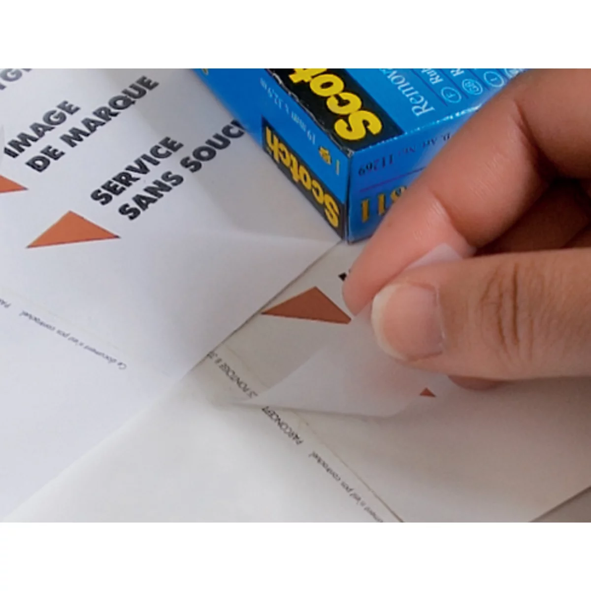 Een hand trekt voorzichtig een label van een vel papier, daarnaast ligt een blauwe Scotch-doos.