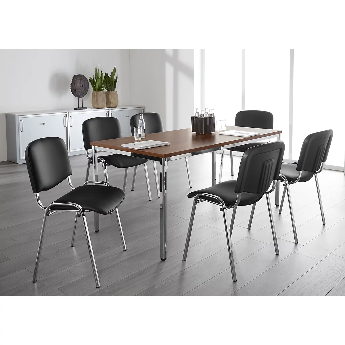Table de conférence avec 6 chaises. Sur la table, de l'eau, un vase et un ordinateur portable. En arrière-plan, des armoires et des plantes.