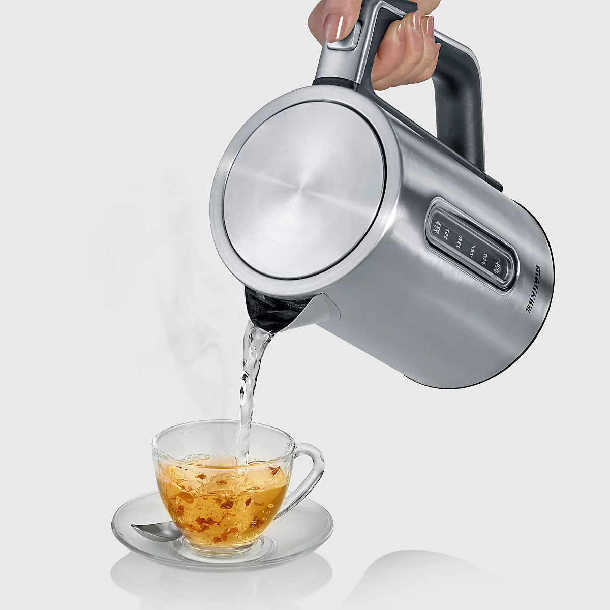 Eine Hand gießt Wasser aus einem silbernen Wasserkocher in eine Teetasse. Dampf steigt auf. Die Tasse steht auf einer Untertasse.