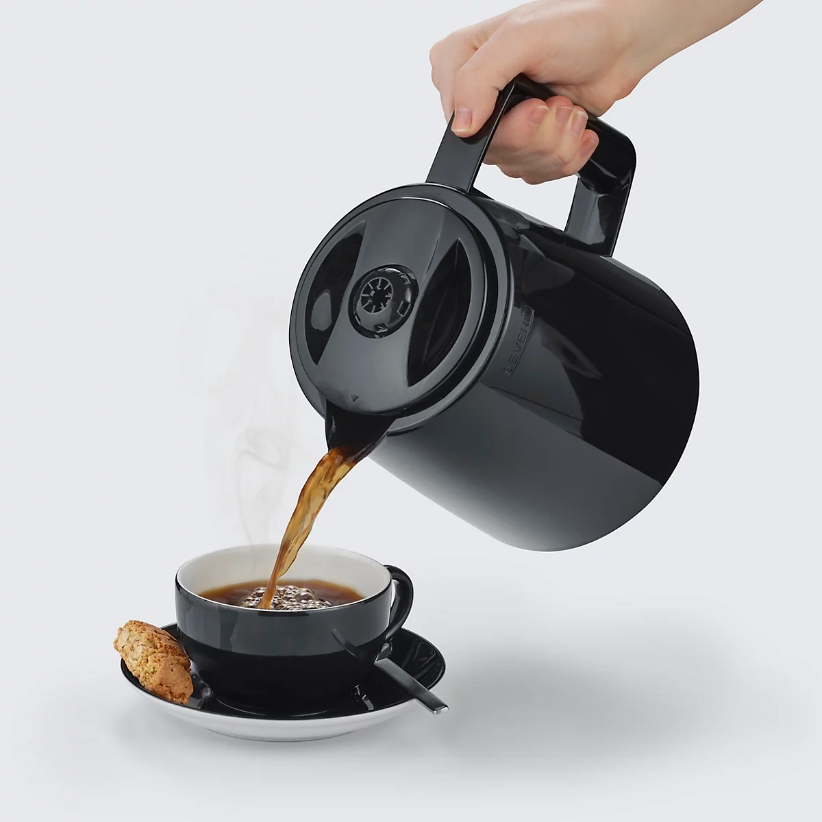 Eine Hand giesst Kaffee aus einer schwarzen Kaffeekanne in eine schwarze Tasse. Auf dem Unterteller liegt ein Keks.