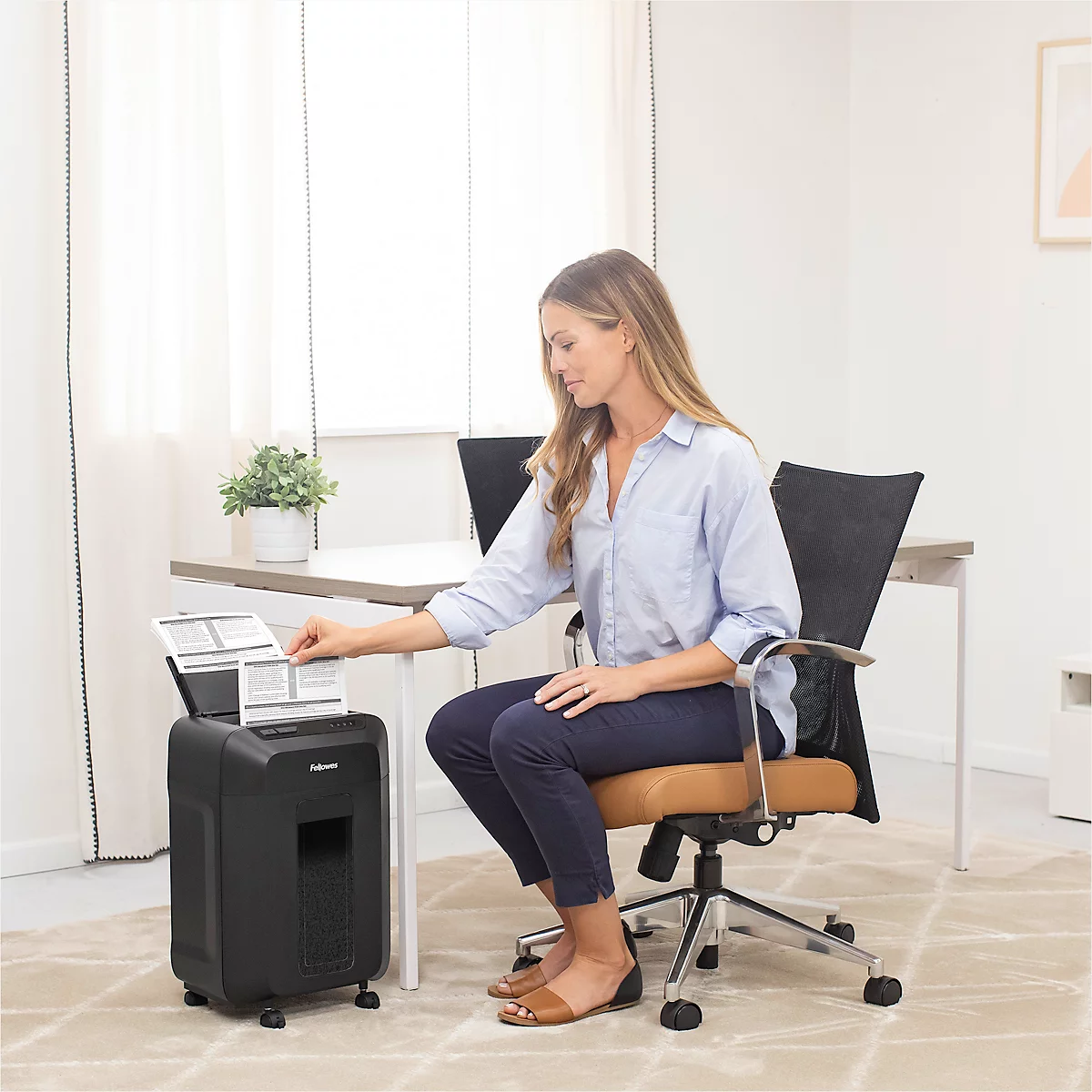 A woman sits at a desk placing documents into a paper shredder. She wears a blue blouse and dark trousers.