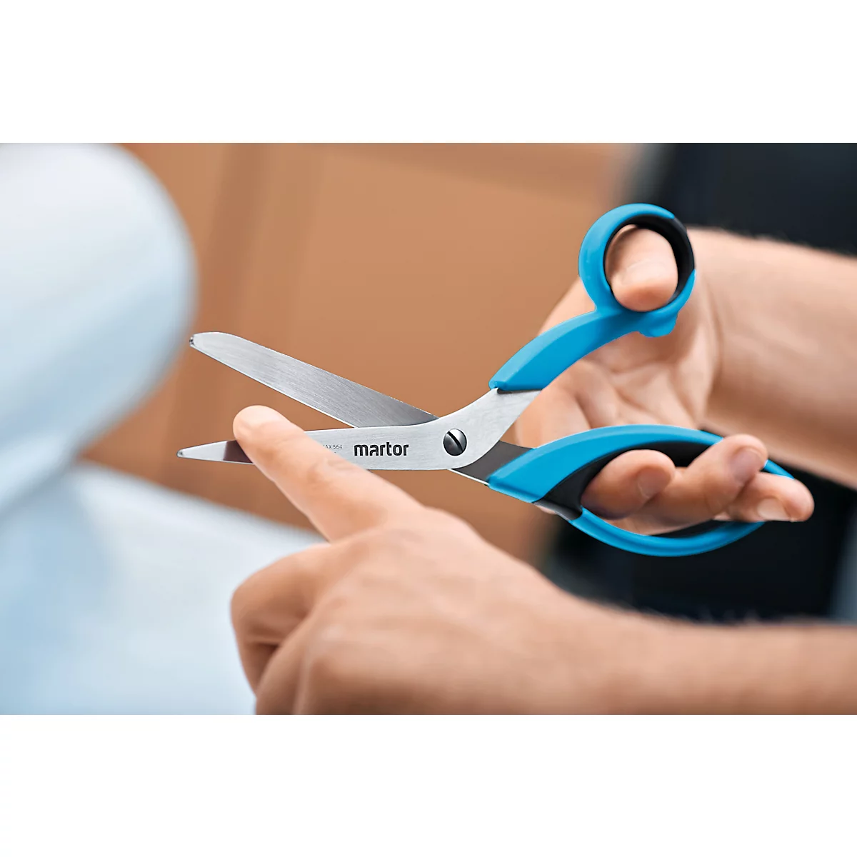 Person hält eine blaue Schere. Die Schere ist geöffnet und die Klinge zeigt nach oben. Der Name "martor" ist auf der Schere zu sehen.