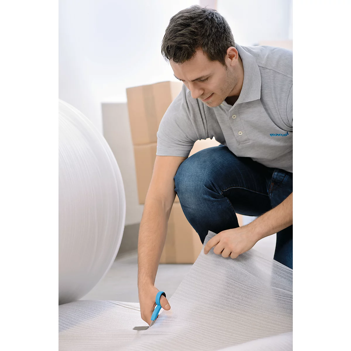 Ein Mann kniet und schneidet Verpackungsmaterial mit einer Schere. Im Hintergrund sind Kartons.