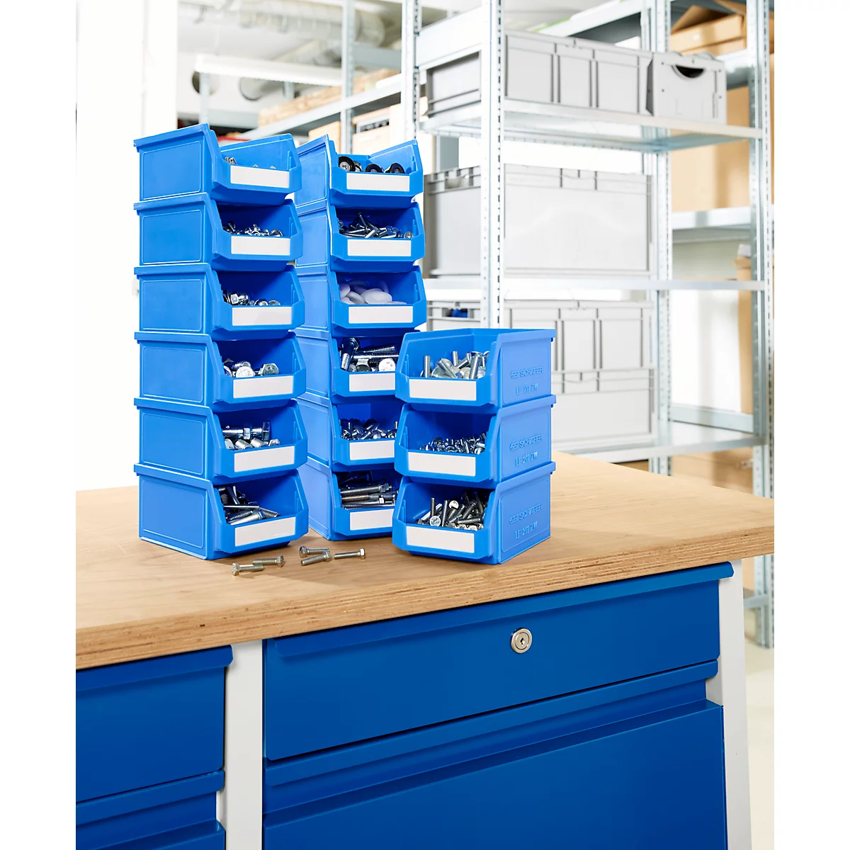 Drei Stapel blauer Sichtlagerkästen mit Kleinteilen stehen auf einer Werkbank in einer Lagerumgebung.