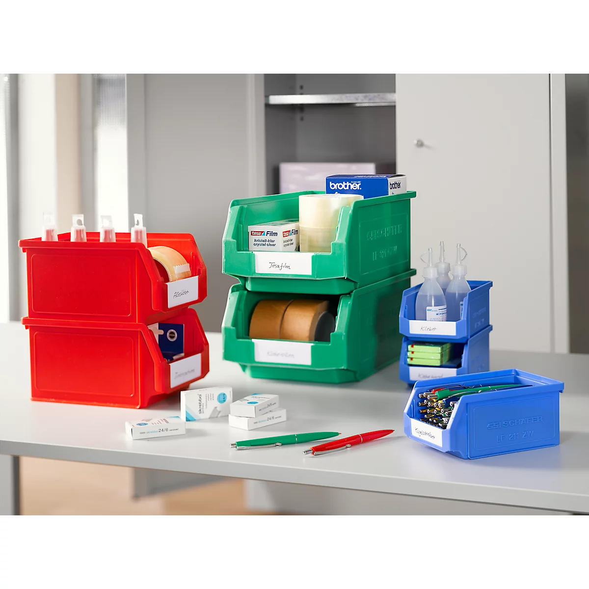 Mehrere rote, grüne und blaue Sichtlagerkästen aus Kunststoff mit Büromaterialien auf einem Tisch.